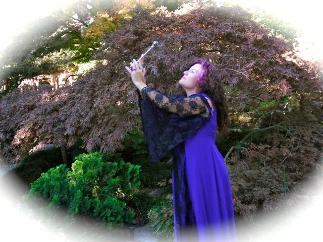 Kim Kelley in a violet gown, raising a Willowroot wand beneath a red maple